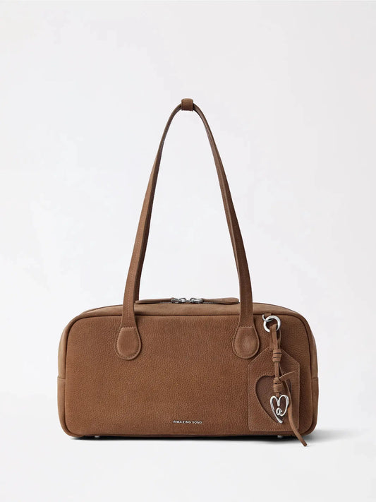 A soft genuine leather travel pillow bag in warm tan resting on a hotel bed beside a folded scarf